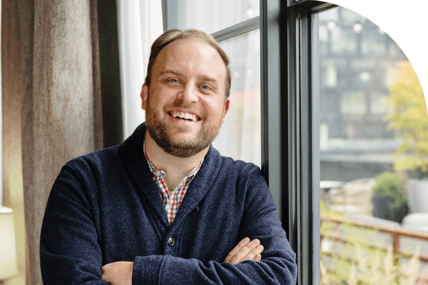 Joshua Lance smiles at camera while leaning against window