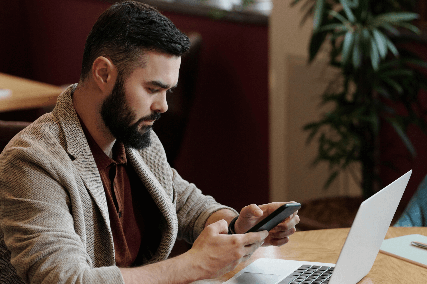 Man sits at table with open laptop while looking at his mobile phone